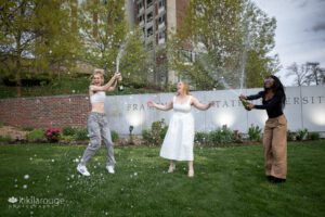 Two friends popping and spraying champagne over their friend in celebration of her graduation in front of Framingham State University sign