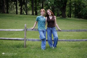 Twin Seniors in jeans leaning on wooden fence is green field