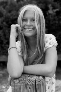 BW portrait of teen blonde girl leaning on wood post laughing with hair blowing across her face