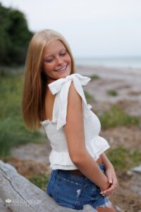 Teen Senior Portrait girl laughing looking down while leaning on large piece of driftwood at beach