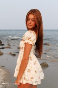 Teen girl in flowery romper with long wet read hair by water's edge at beach looking down