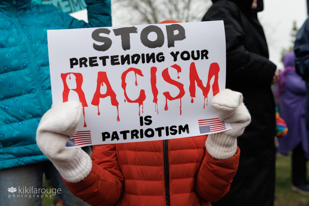 Young person hidden behind sign saying Stop Pretending your Racism is Patriotism with two upside down flags.