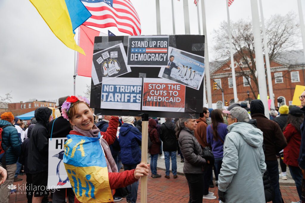 Woman wrapped in Ukrainian flag holding up signs at Hands Off rally in Salem MA