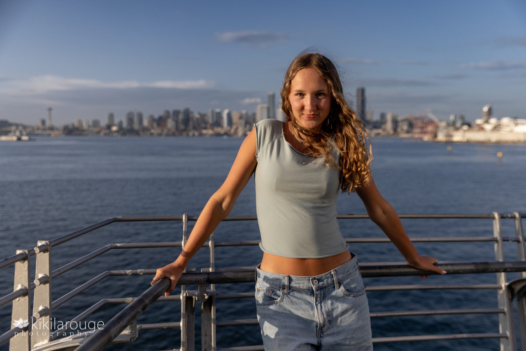 Senior girl in jeans and green crop top leaning on railing smiling with the Seattle skyline blurred out in the backdrop