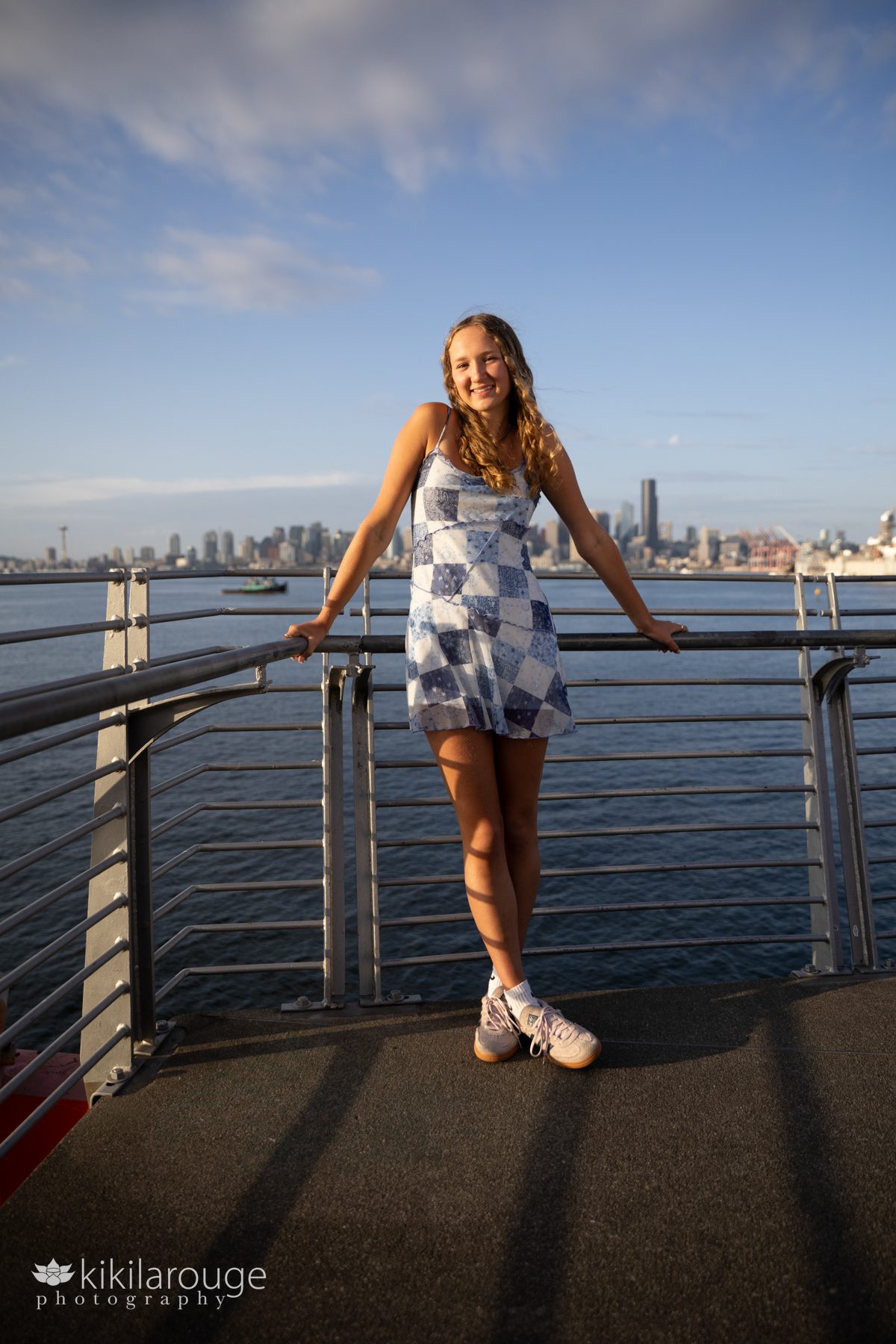 Senior girl in white and blue mini dress and sneakers leaning on railing smiling with the Seattle skyline blurred out in the backdrop