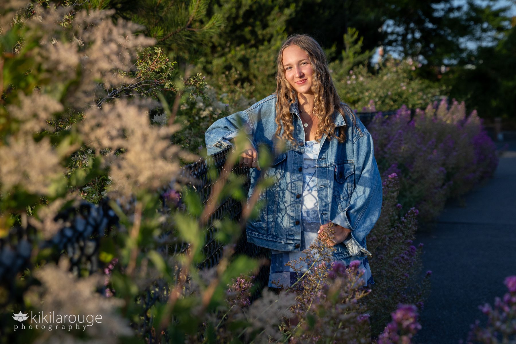 Teen girl in blue and white dress with denim jacket leaning on barbed wire fence surrounded by June flowers and flora in bloom West Seattle