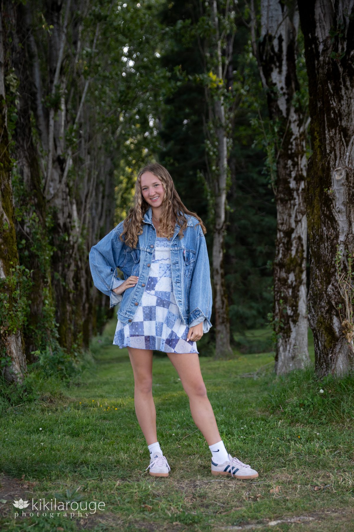 Teen girl in blue and white dress with denim jacket standing with hand on hip in perfect line of PNW trees on either side