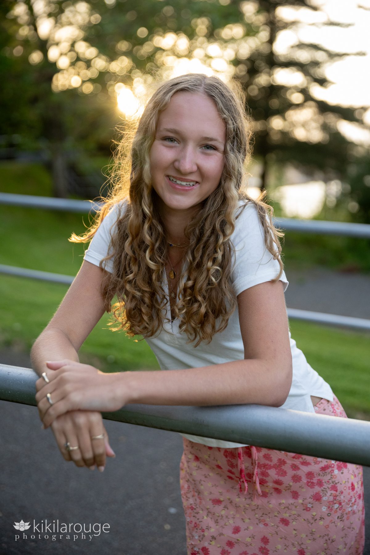 Pretty blond Senior girl leaning on metal railing with sun flare lighting her from behind white shirt and floral pink skirt
