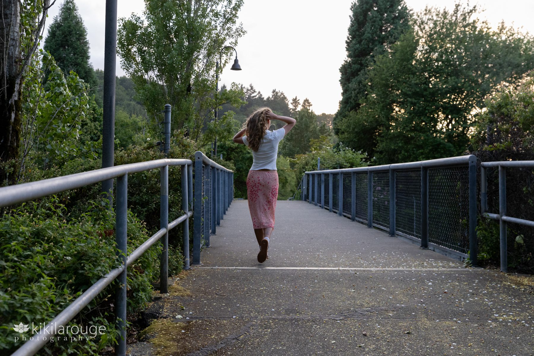 Teen girl with hands in hair walking away on bridge with metal railings wearing long pick floral skirt and white top