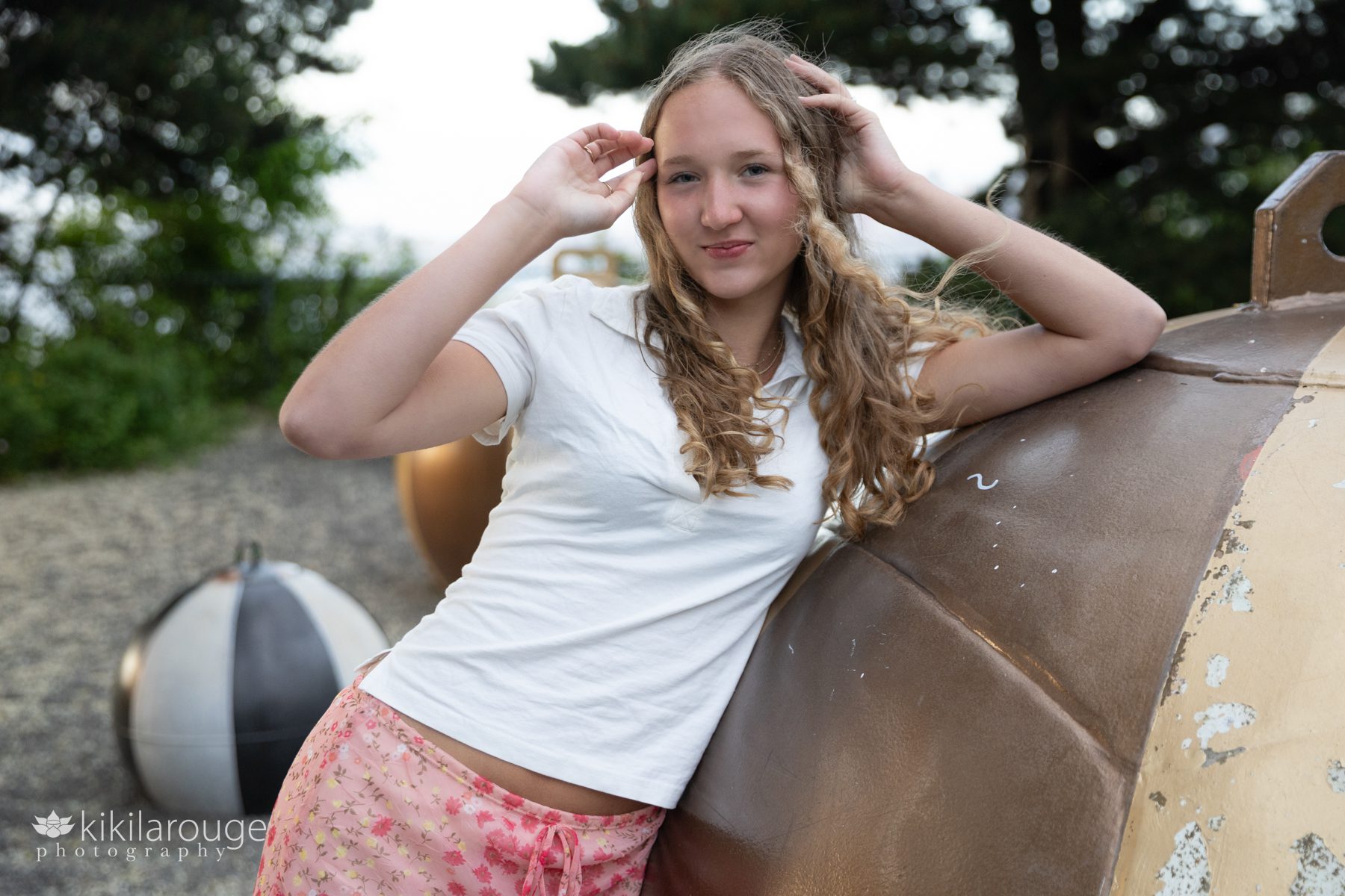 Teen girl leaning on large metallic nautical structure in white top and floral pink skirt