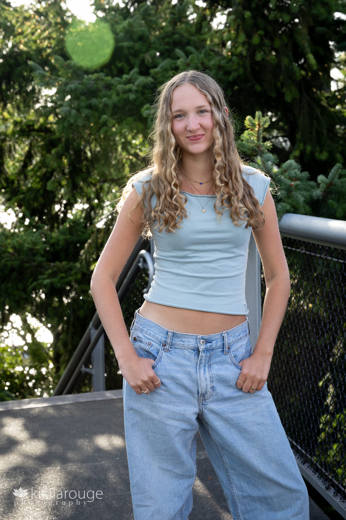 High School Senior with long curly blonde hair in light green top standing with hands in jeans and PNW flora in the back drop