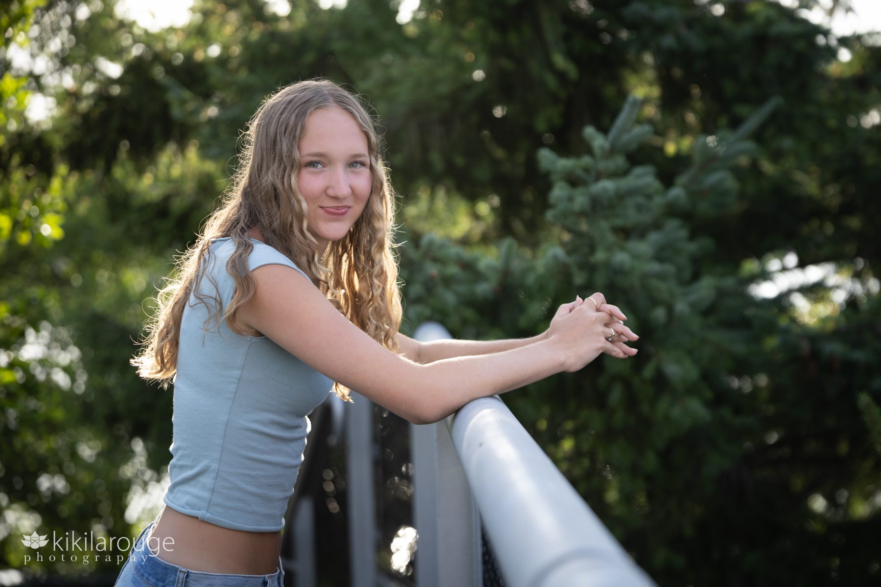 High School Senior with long curly blonde hair in light green top and jeans leaning on metal rail with PNW flora in the back drop