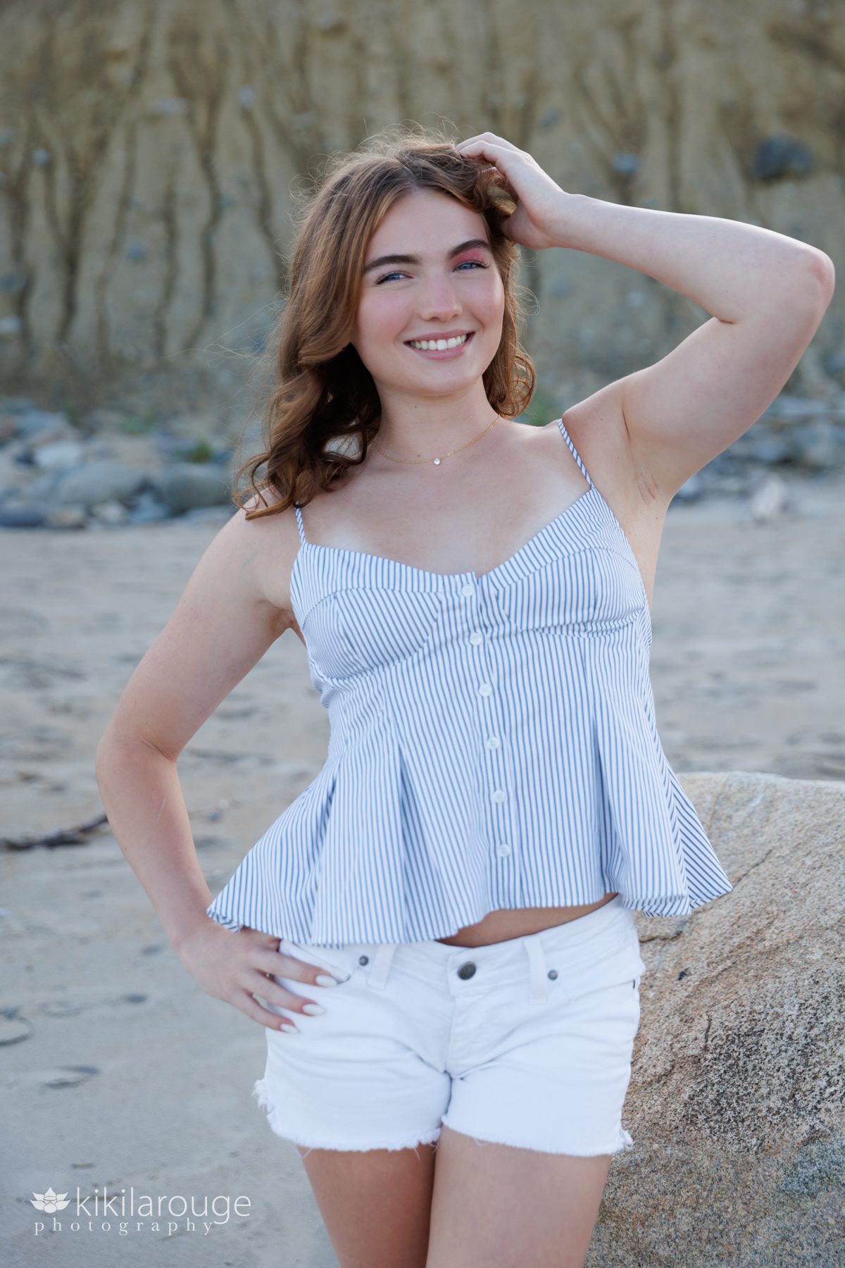 Senior girl with reddish shoulder length hair in striped tank top and white shorts leaning on large rock at beach hand in hair smiling