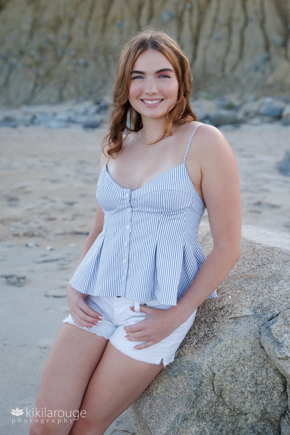 Senior girl with reddish shoulder length hair in striped tank top and white shorts leaning on large rock at beach