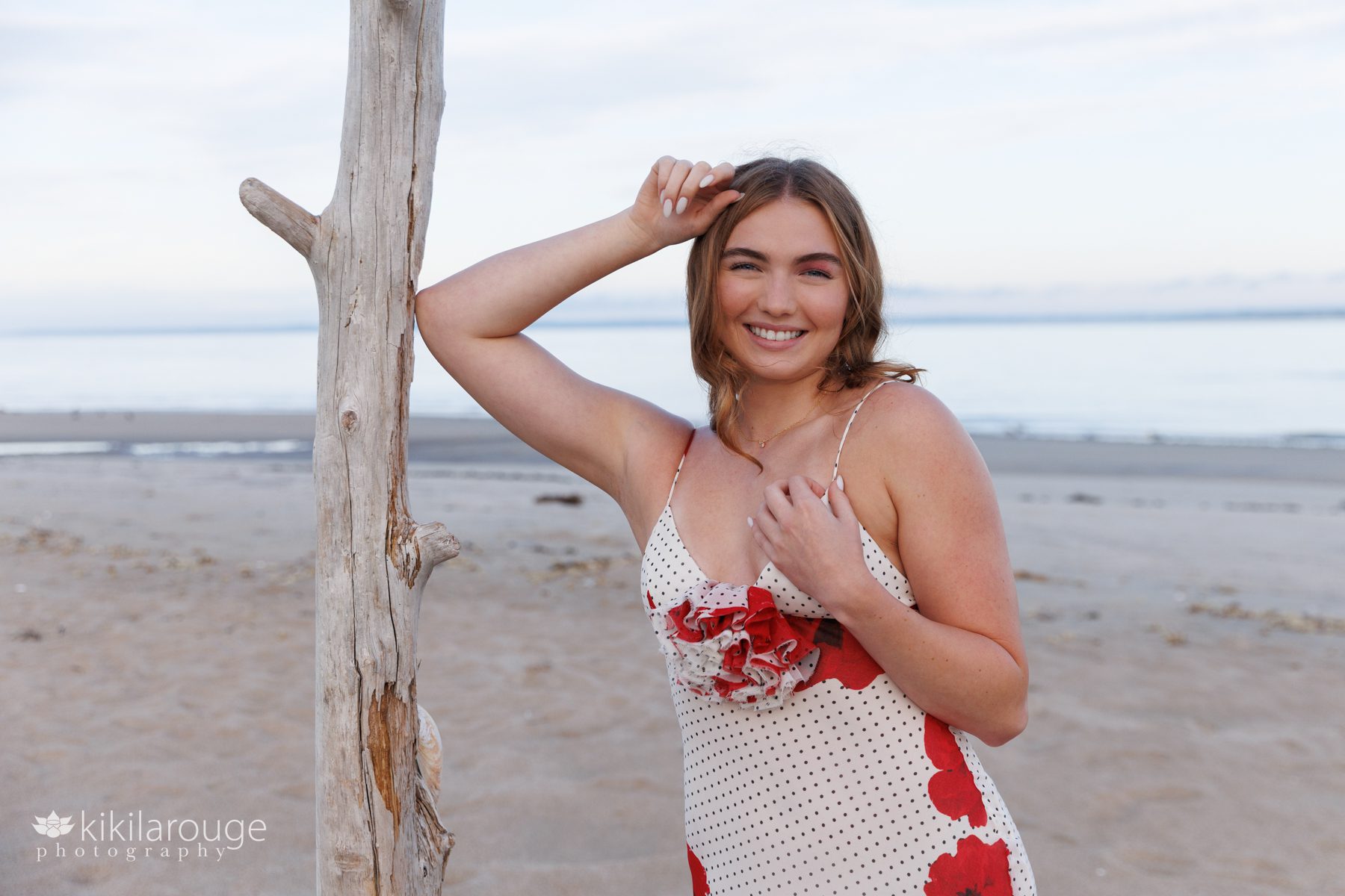 Girl in long floral dress with black polka dots leaning on a piece of bleached out driftwood with ocean in backdrop