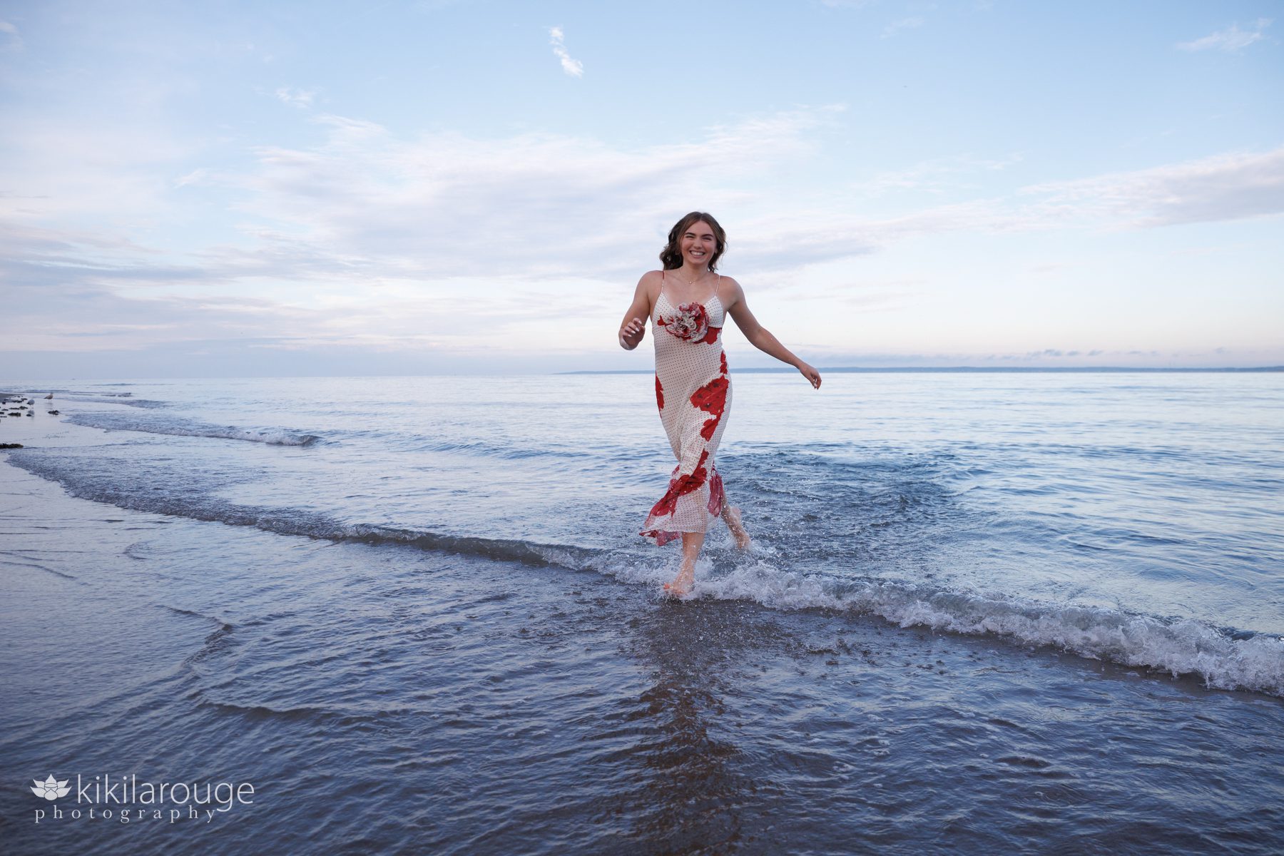 Girl running along shoreline with reflection in long floral dress with black polka dots smiling and water splashing