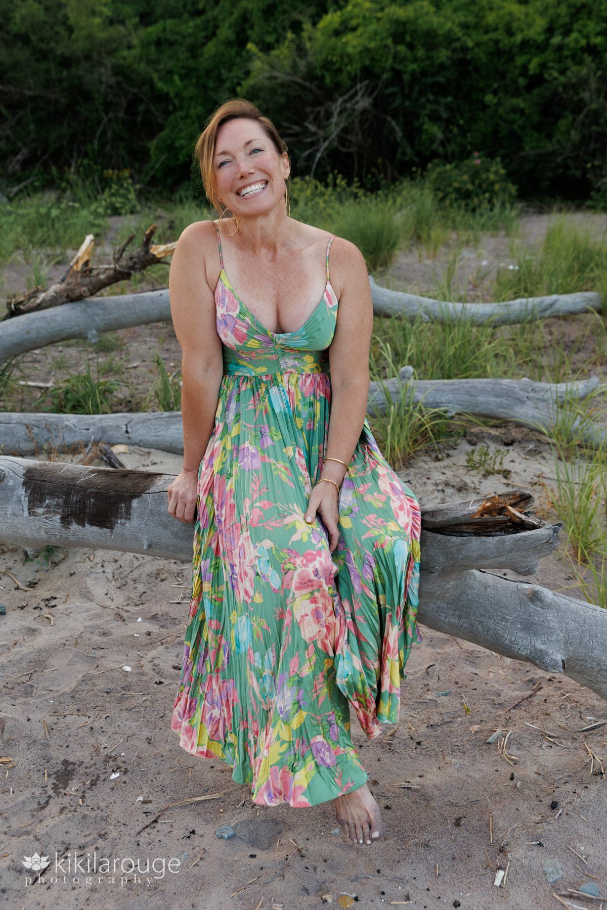 Woman with big smile in floral green summer dress sitting on driftwood at Sandy Point beach with green shrub backdrop and purple sand