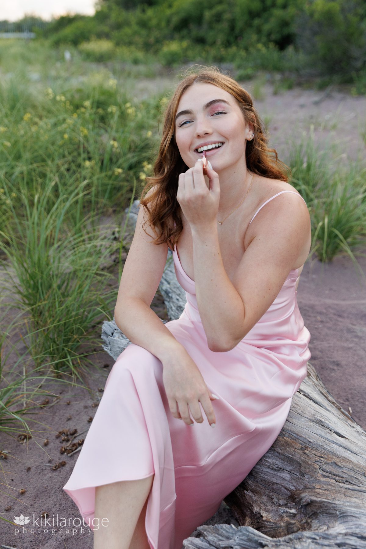 Girl applying lipgloss at photo shoot while sitting on driftwood surrounded by dunes and purple sand