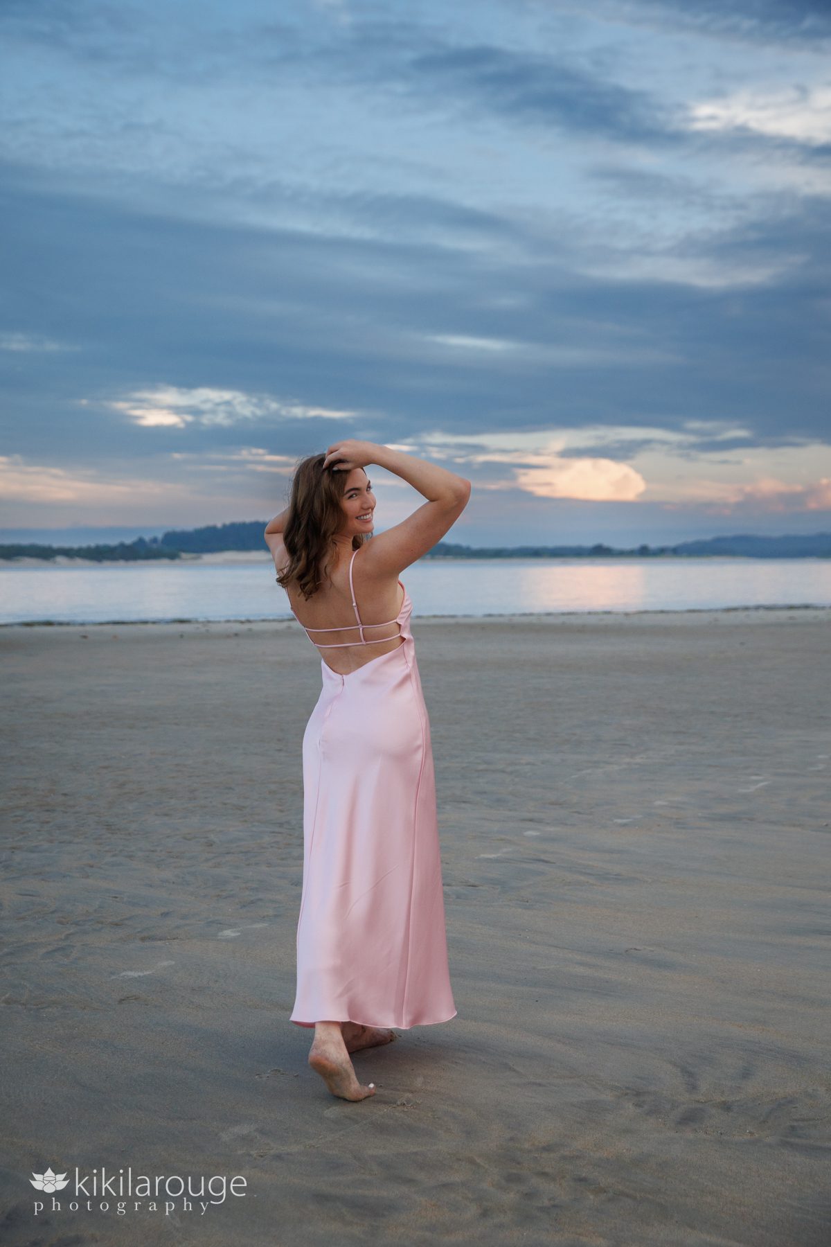 Teen girl walking away on wide open beach looking back over her shoulder with her hands in her hair in a pink satin dress