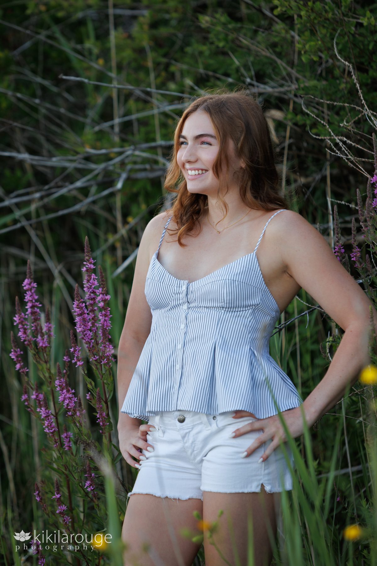 Senior girl with reddish shoulder length hair in striped tank top and white shorts standing in beach dunes with purple and yellow flowers