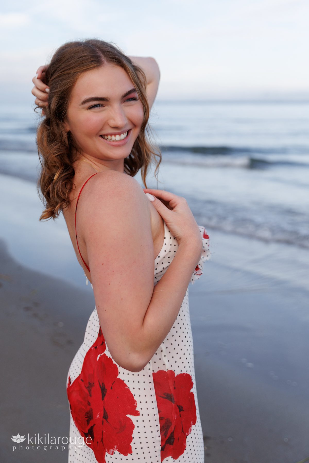 Teen girl in floral dress with large red flowers and polka dots with hand on décolletage and other in her hair with beach waves in backdrop