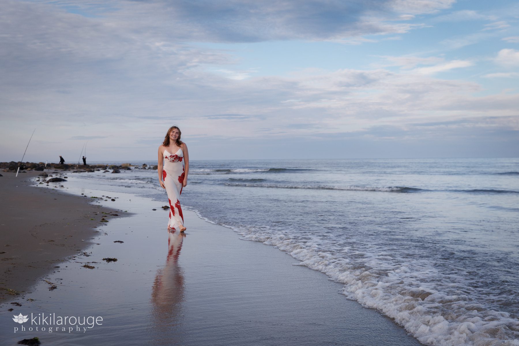 Girl walking along shoreline with reflection in long floral dress with black polka dots and two fishermen in the backdrop