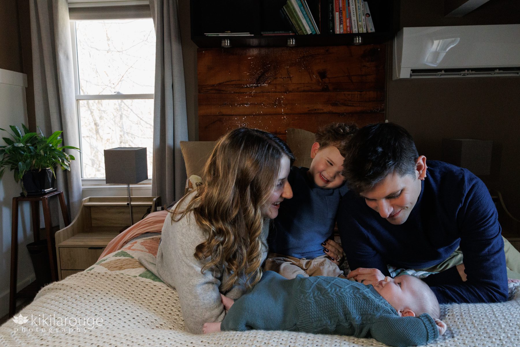 Mom, Dad and three year old smiling and looking down at newborn baby all laying on bed with wood back drop and book shelves 