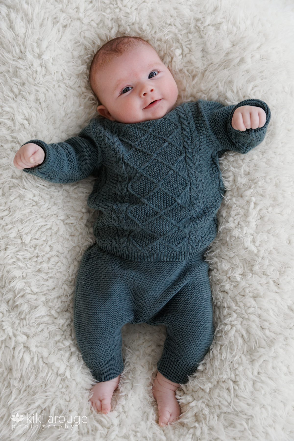 Smiling adorable two month old baby girl laying on soft fluffy blanket dressed in a forest green cable night sweater and pants