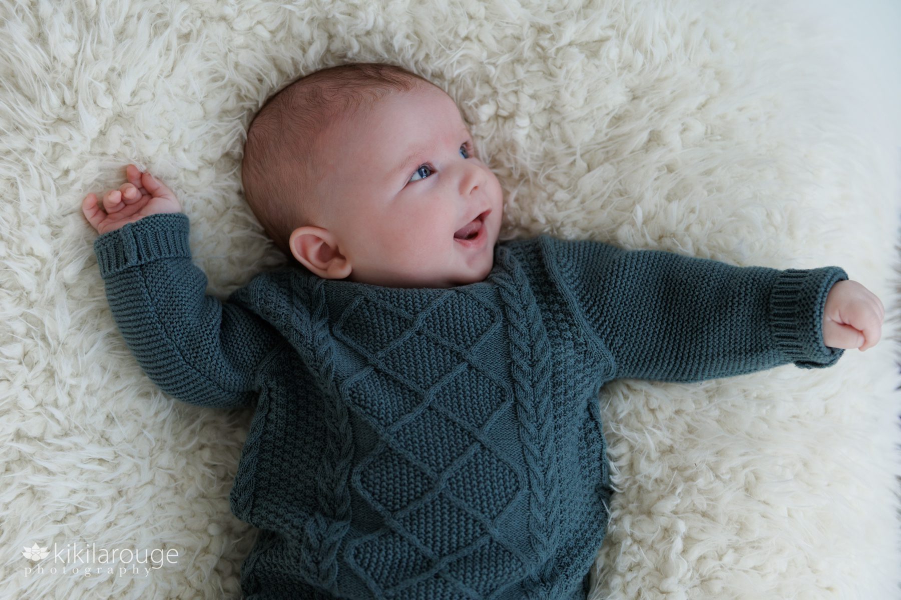 Two month old baby girl in green sweater on fluffy white blanket laughing and smiling at parent who is off to the right
