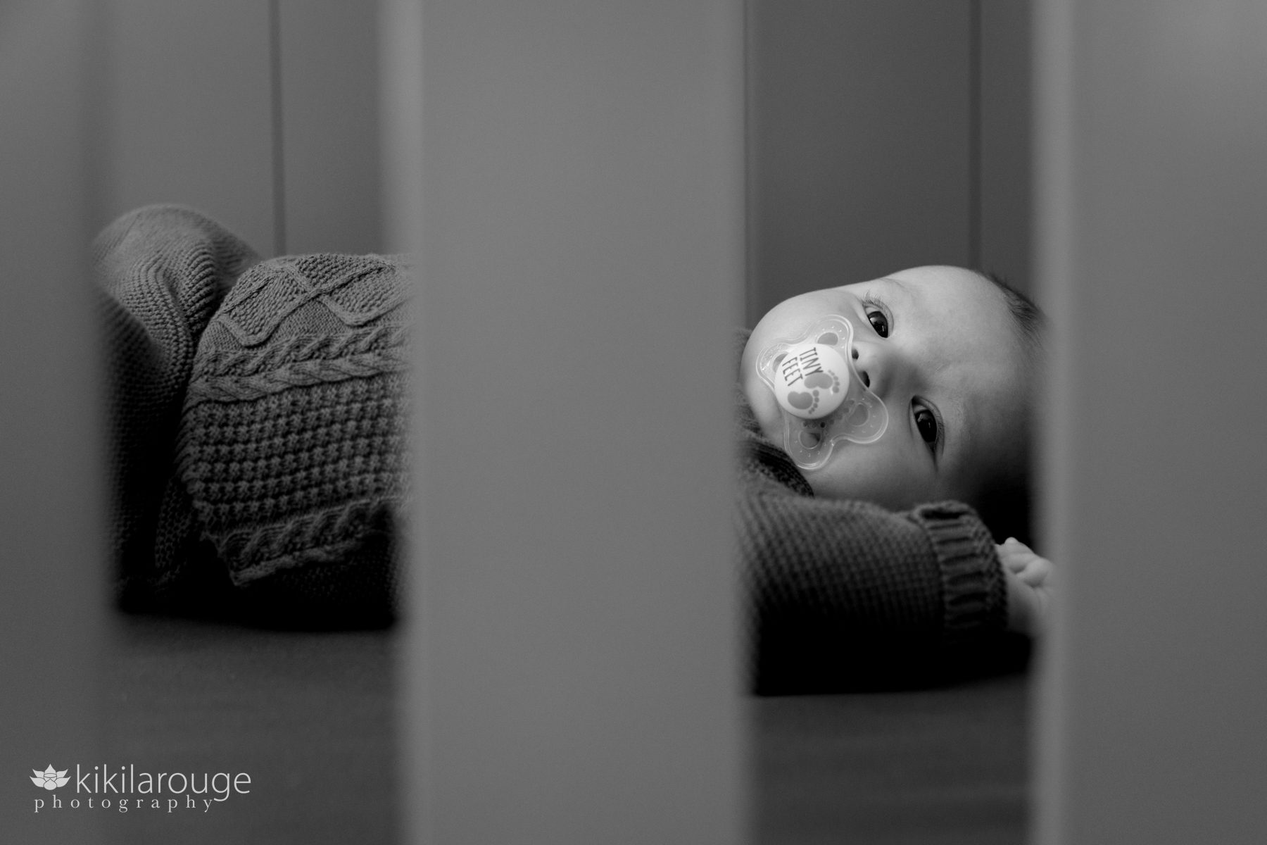 Two month old baby laying in crib with pacifier that says Tiny Feet