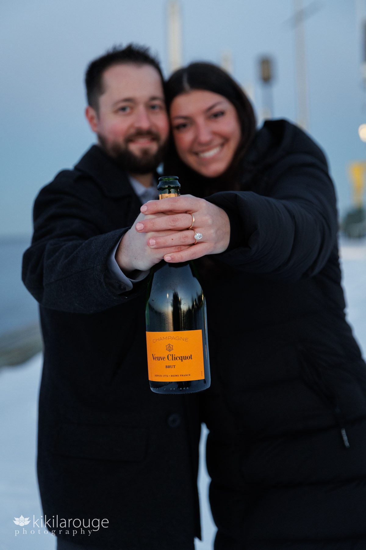 Cute newly engaged couple in black winter coats smiling holding a bottle of Veuve Cliquot champagne and her engagement ring in focus