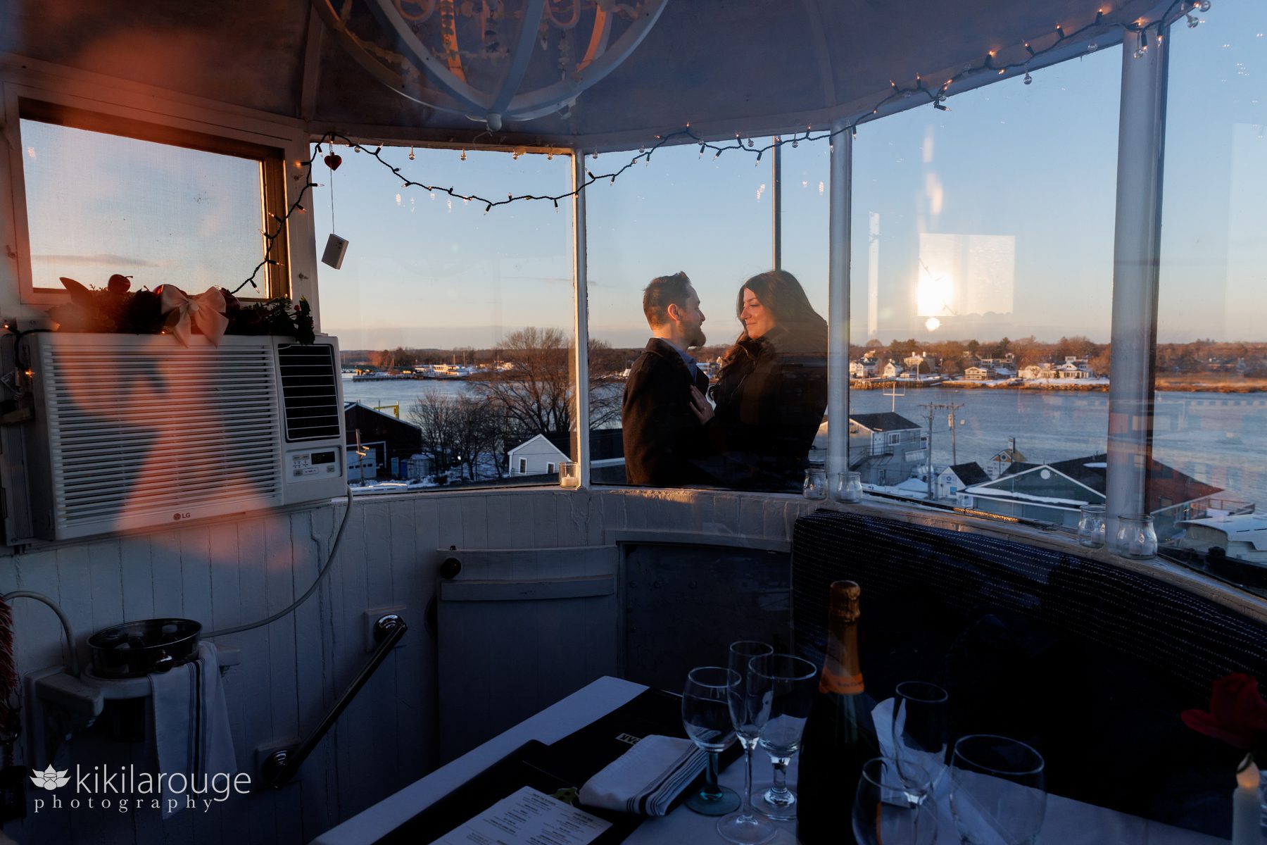 Couple on the outside of lighthouse smiling with NBPT waterfront in the back drop and set table for dinner with bottle of Veuve Cliquot champagne
