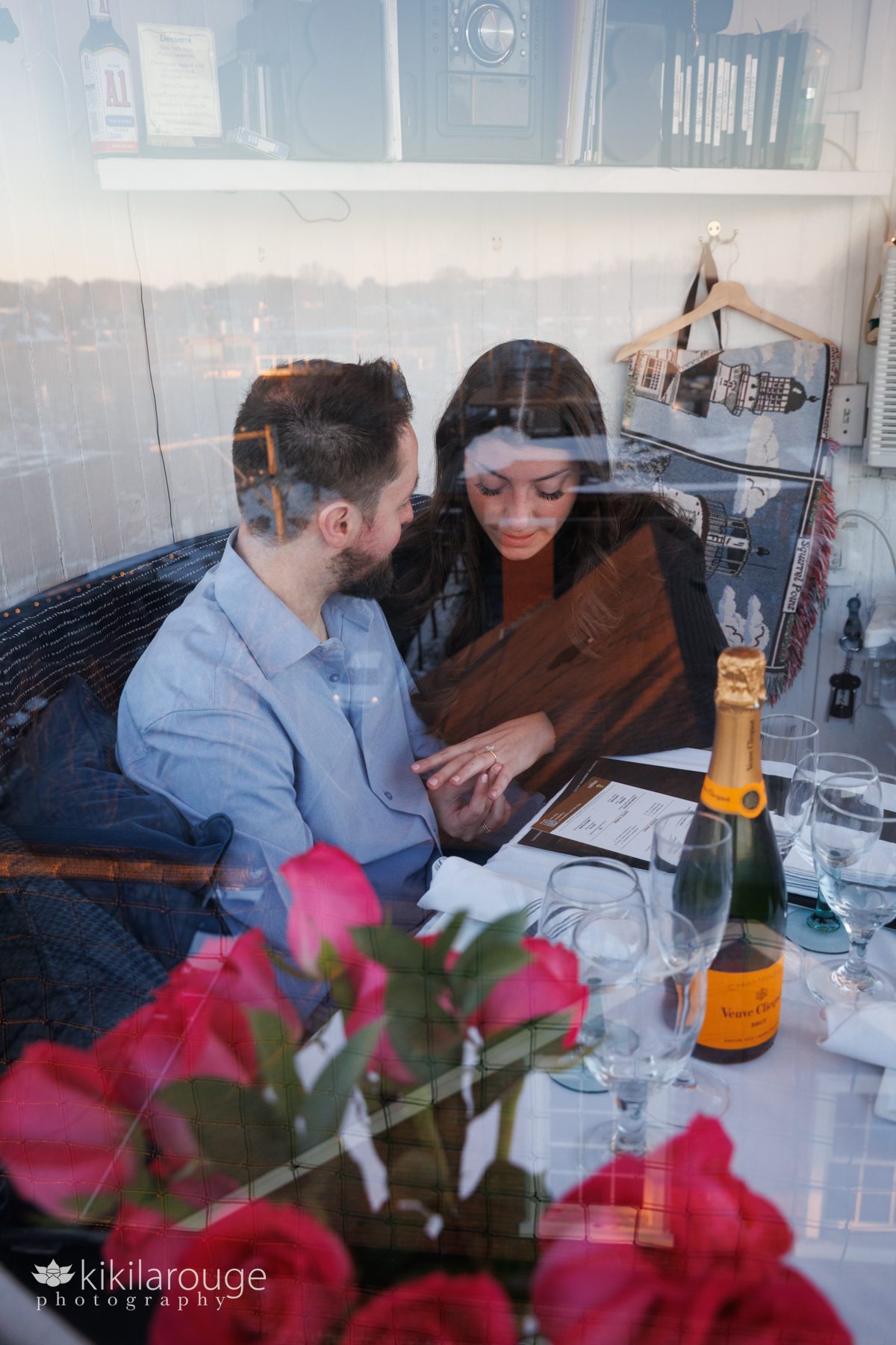Couple inside lighthouse looking at new engagement ring with bottle of Veuve and pink flowers on the table shot through the lighthouse glass so reflections
