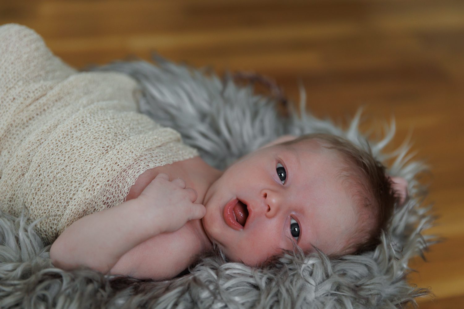 Newborn in nude cheesecloth wrap laying on gray blanket with big open eyes and mouth
