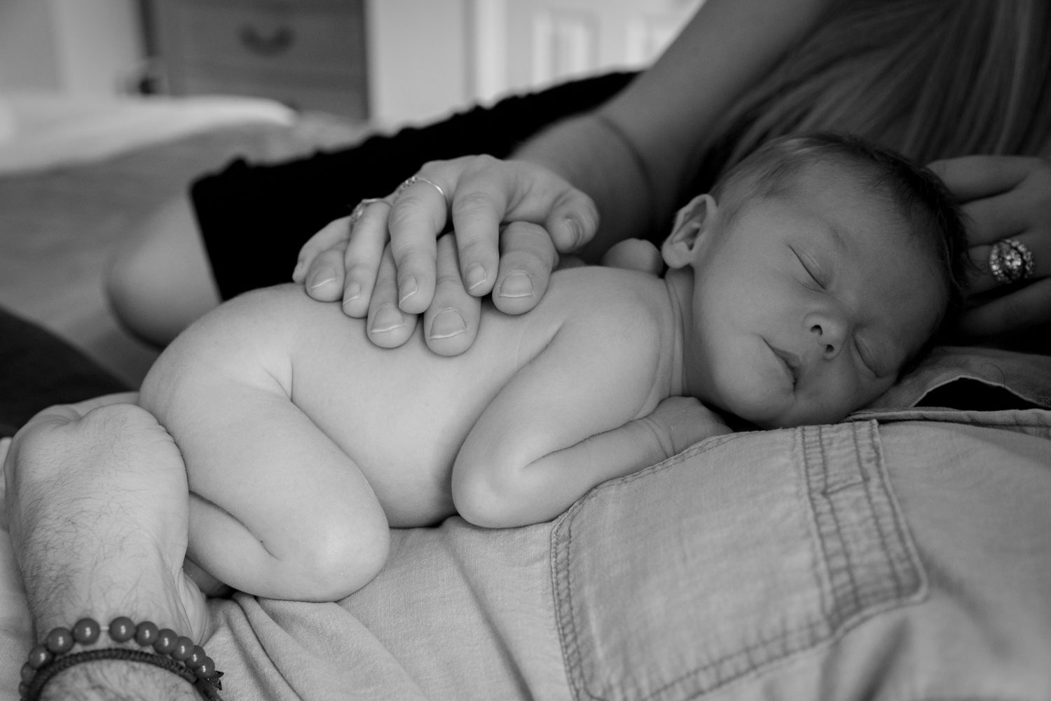 Naked newborn baby sleeping on Dad's chest with Mom and Dad's hand on his back