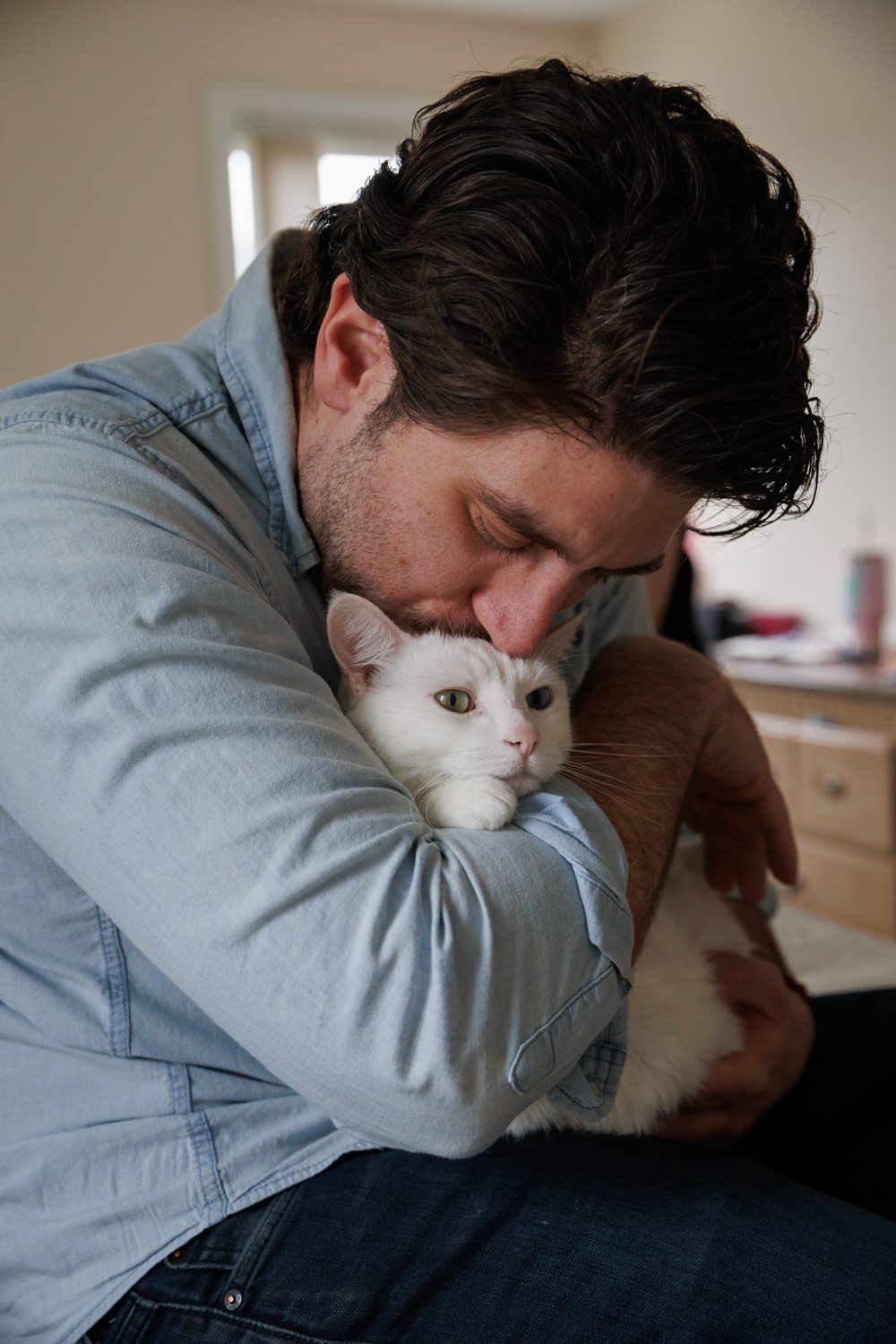 Man in jeans and light blue shirt snuggling with a white cat with one green and one blue eye