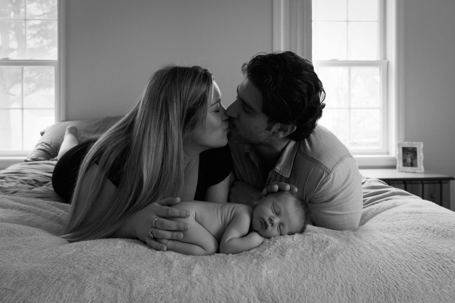 Couple laying on bed kissing with sleeping newborn below them each with their hand on him