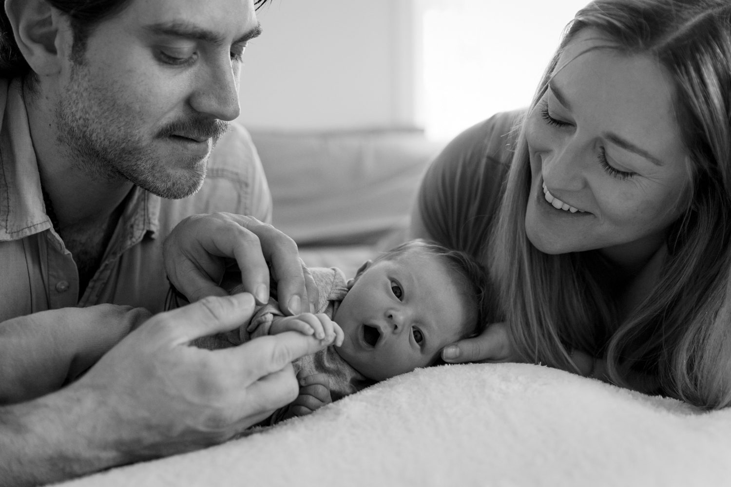 Mom and Dad smiling looking down on baby boy who is wide eyed looking into the camera while holding Dad's finger with his little hand