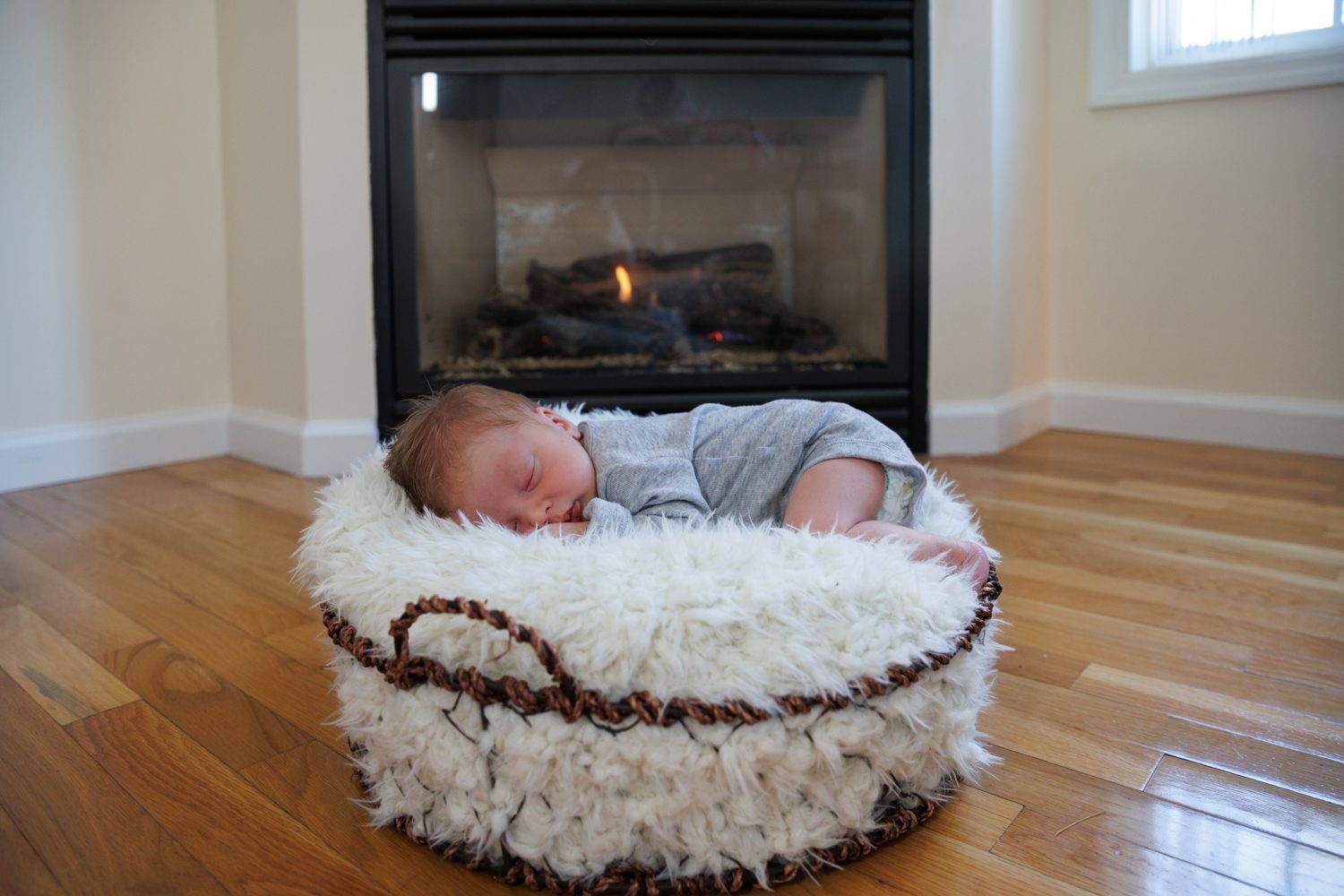 newborn sleeping in wire basket with fluffy blanket in front of a gas fireplace