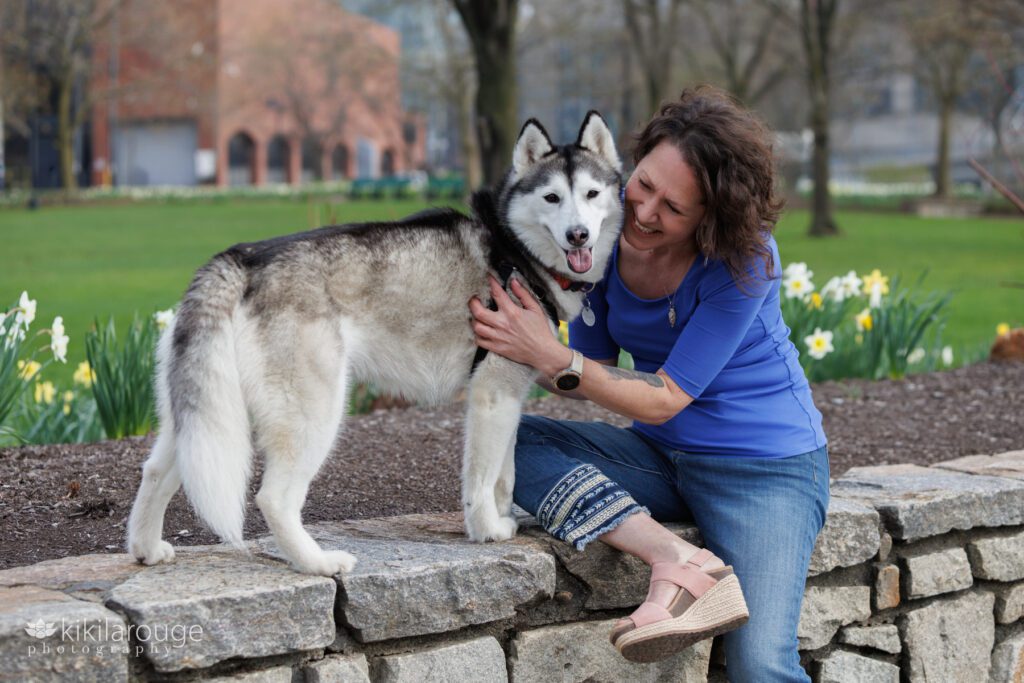 Woman in blue top and embroidered jeans sitting on light stone wall holding and smiling at her husky dog who is looking into the camera