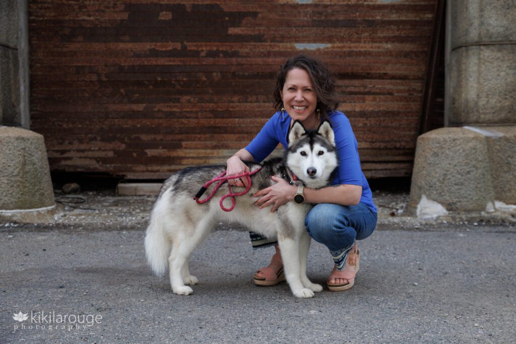Woman in royal blue shirt with embroidered jeans hugging a husky in front of rusted large door