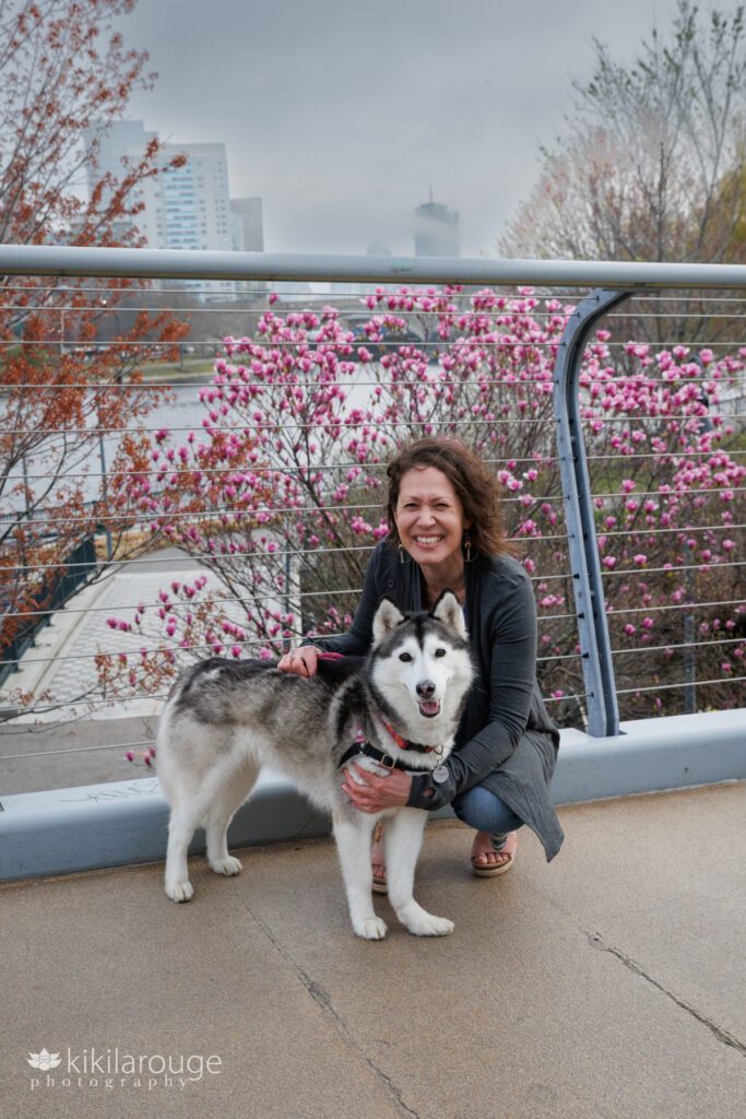 Dog Mom in jeans and long gray sweater holding cute husky dog on the North Bridge Park bridge with magnolias in bloom and the iconic Prudential building in the fog in the backdrop