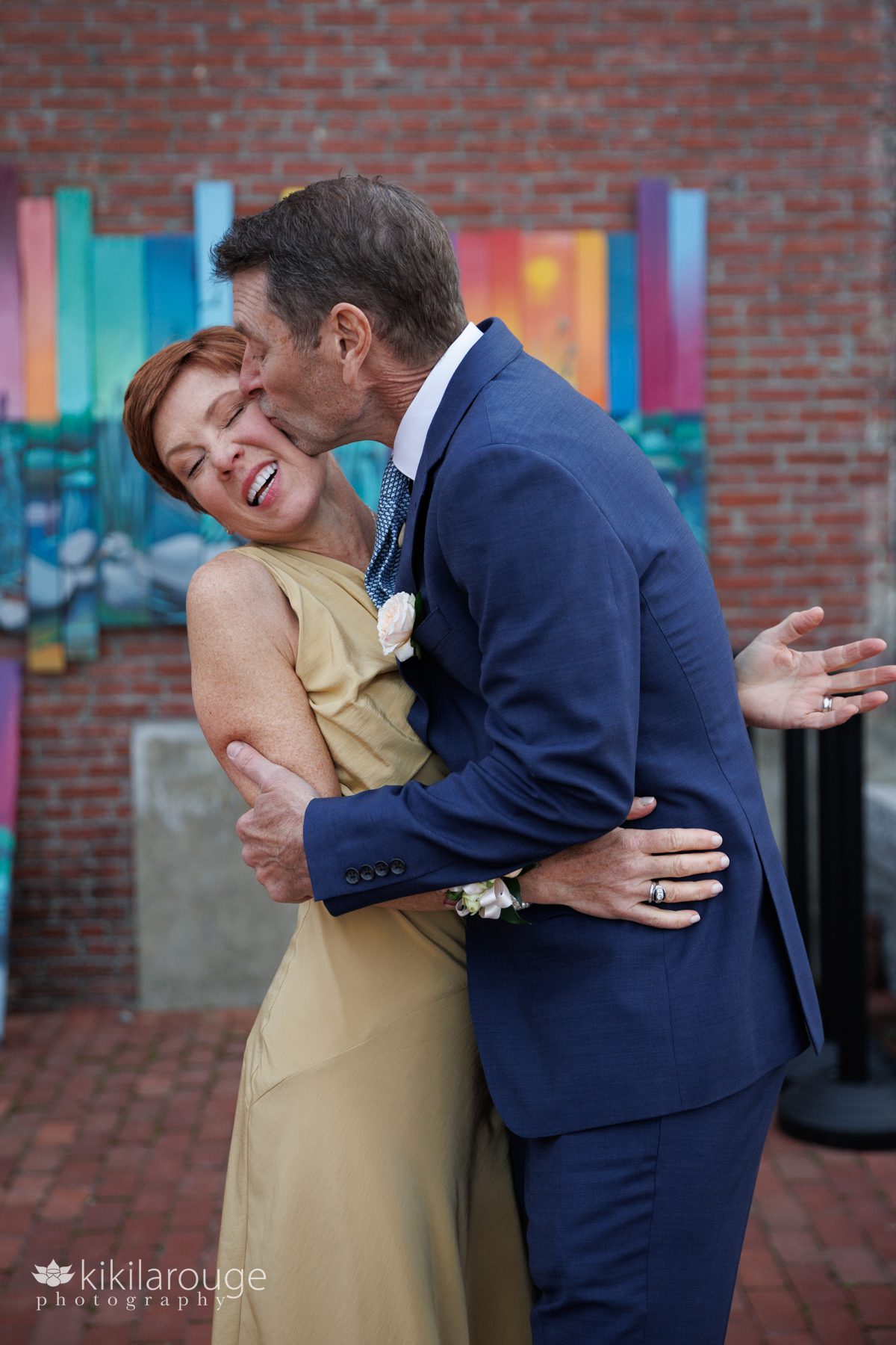 Man in blue suit giving a big kiss on the cheek to a woman smiling and leaning back in front of brick building with colorful mural
