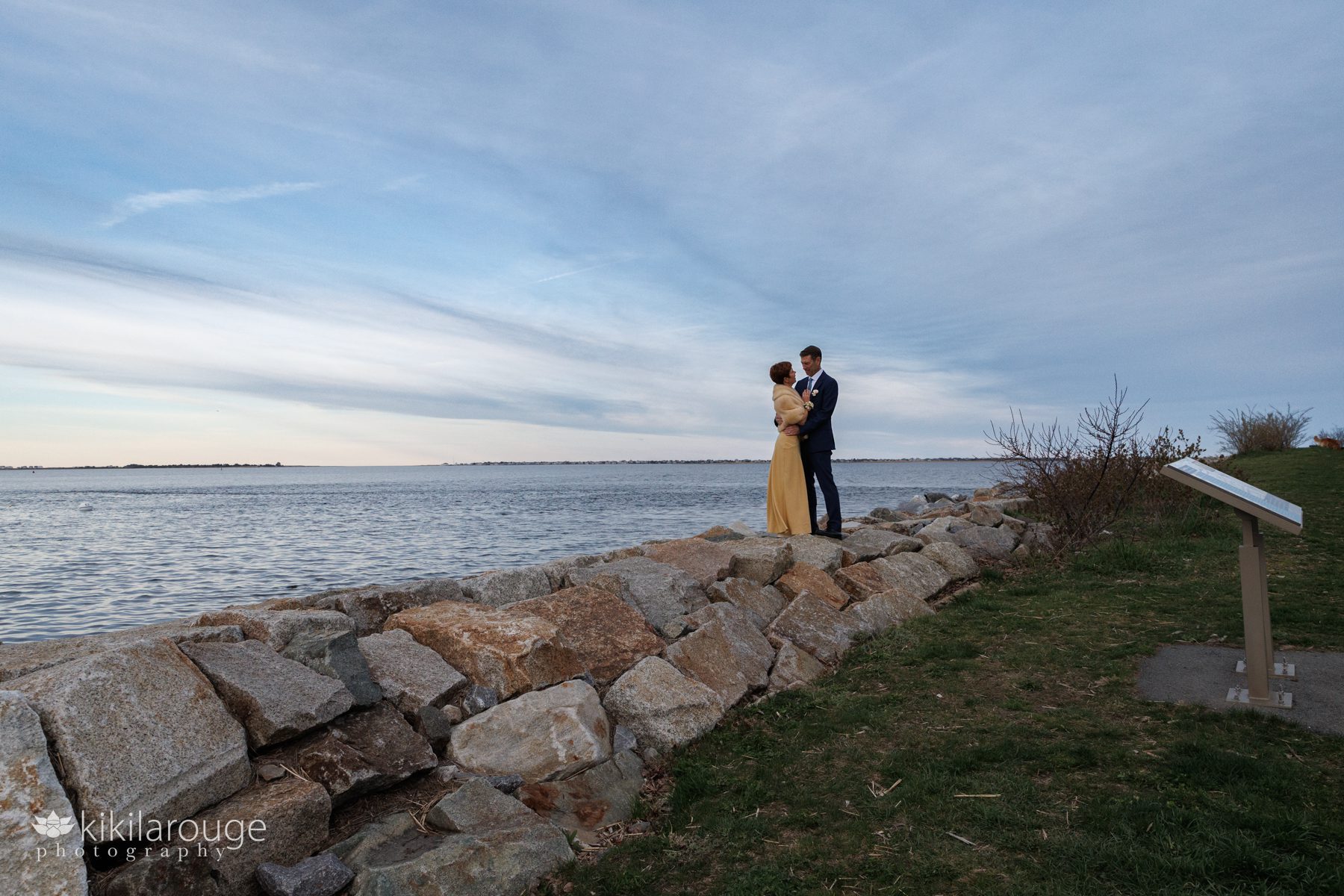 Couple on stone breakwater at sunset with views of the water and Plum Island in the backgroud