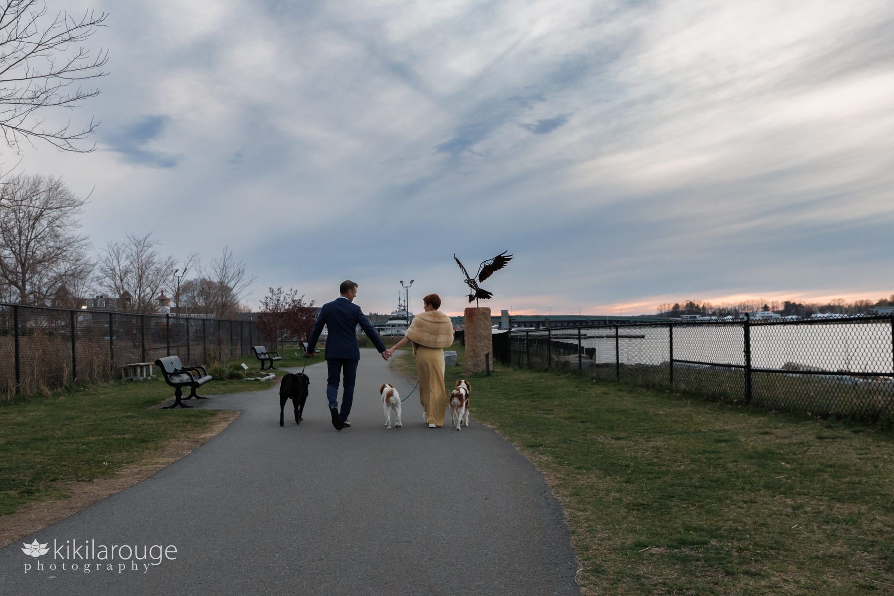 Couple holding hands walking with their three dogs as sunset with the silhouette of a large osprey statue and the Merrimac River