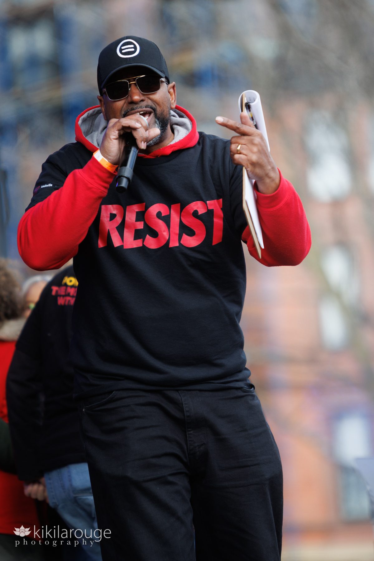 emcee of the No Kings Boston 3.0 Rally in a red sweatshirt and black tee on top with red RESIST speaking in the microphone with clipboard
