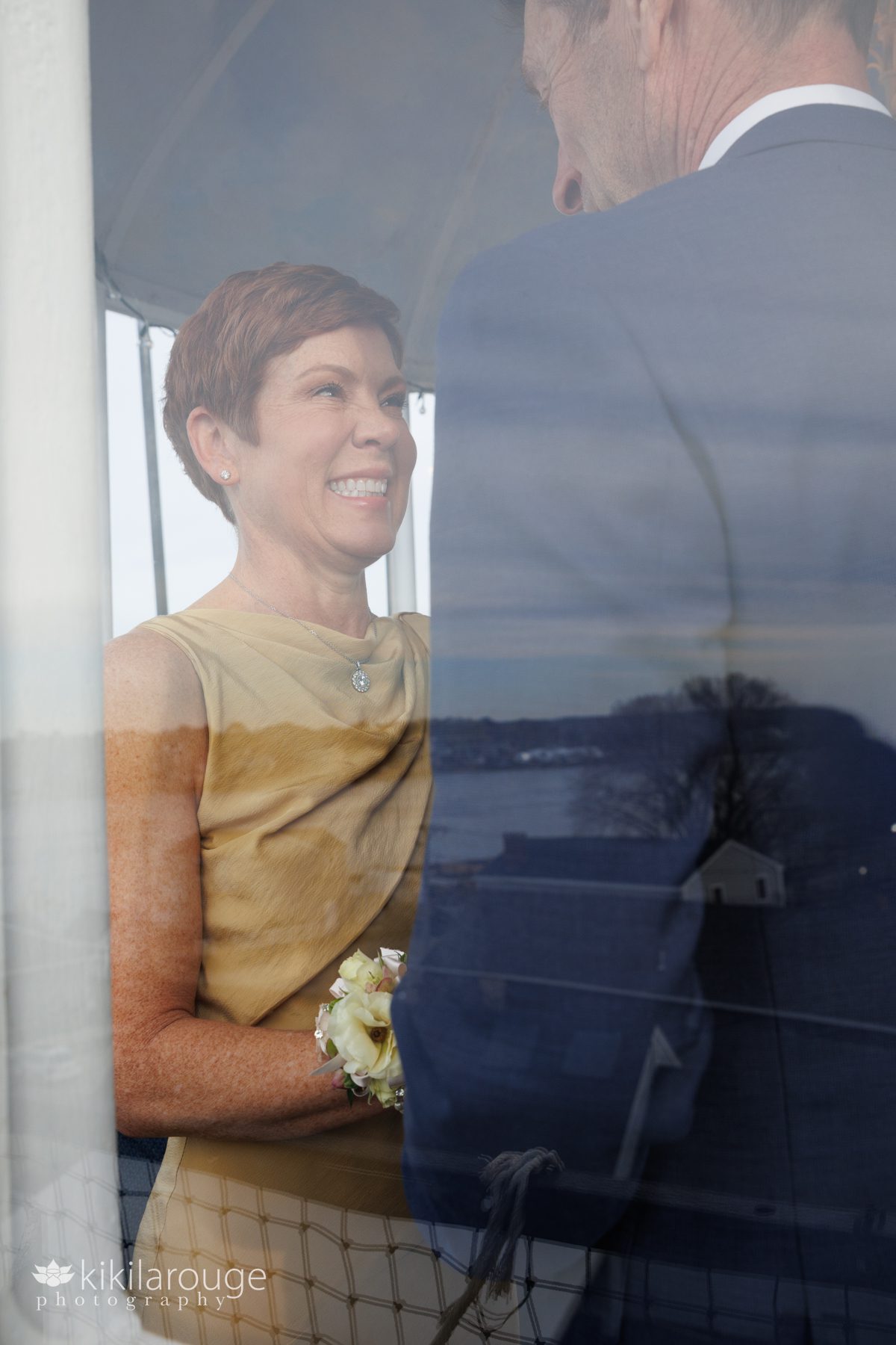 Woman in golden dress smiling at man in blue suit while getting married atop the Newburyport Lighthouse. 