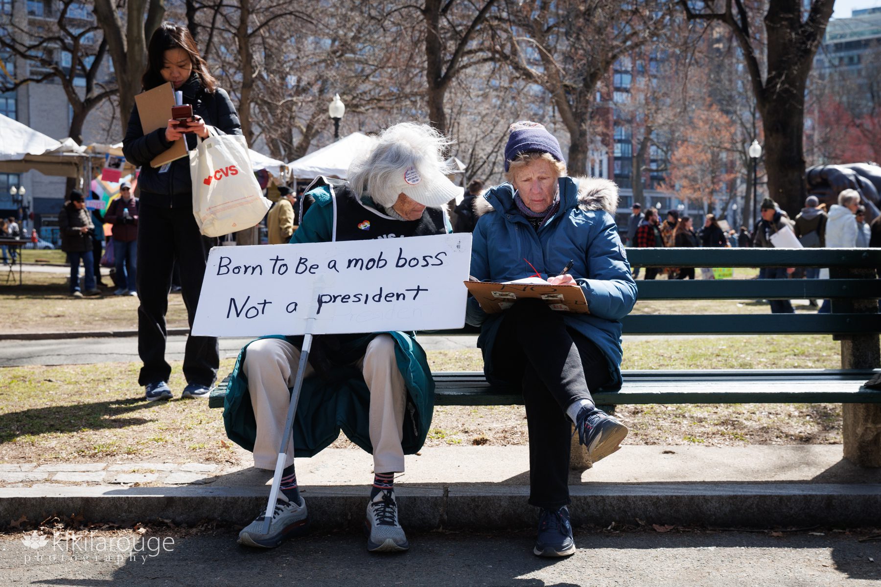 Two older women on Boston Common park bench with protests signs at the No Kings 3.0 protest