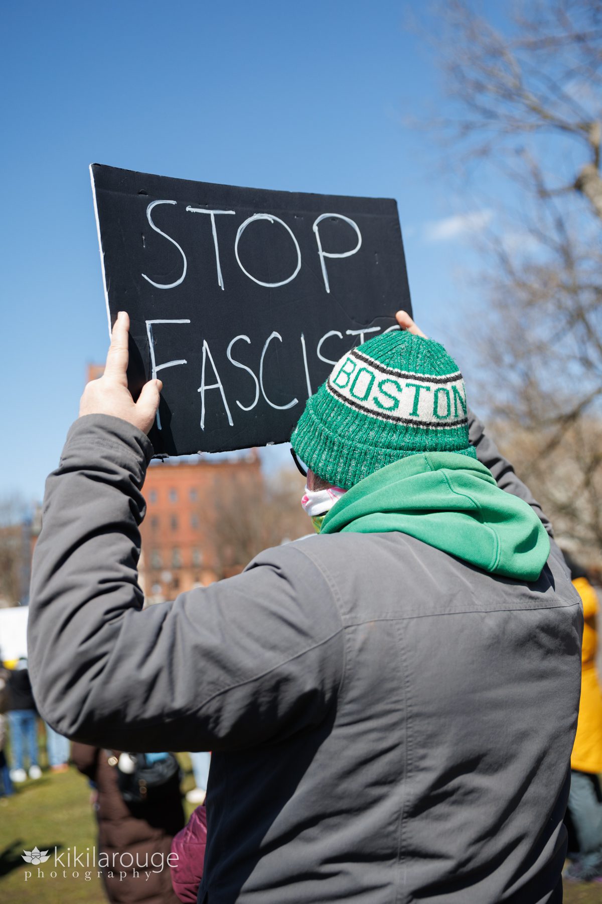 Guy in green Boston winter hat holding up a black sign that says STOP FASCISTS
