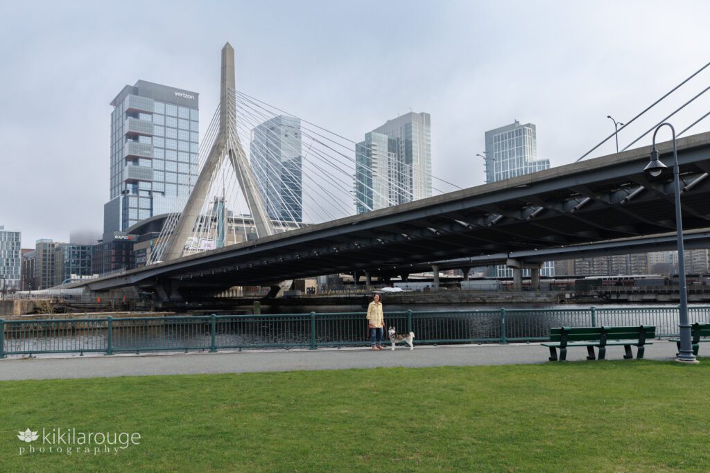 Wide out negative space photo of woman in yellow floral vintage coat under the Zakim Bridge next to Charles River with the Boston Garden in the backdrop